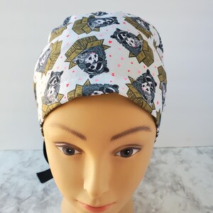 May include: A white surgical scrub cap featuring a repeating pattern of raccoons in cardboard boxes and small pink hearts. The raccoons are depicted in shades of gray, black, and white, with the boxes in shades of brown and tan.