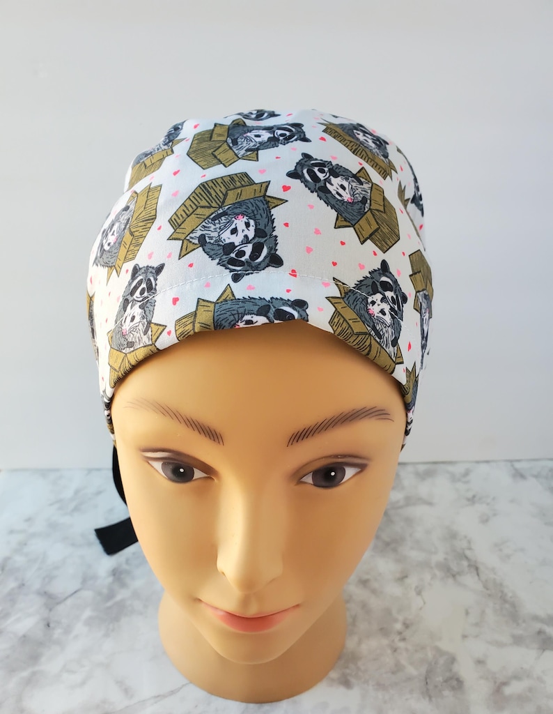 May include: A white surgical scrub cap featuring a repeating pattern of raccoons in cardboard boxes and small pink hearts. The raccoons are depicted in shades of gray, black, and white, with the boxes in shades of brown and tan.