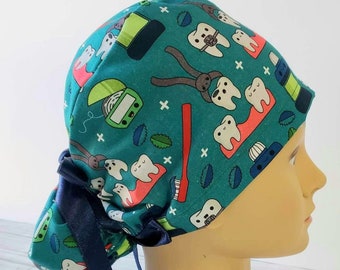 May include: A teal scrub cap featuring a repeating pattern of cartoon teeth, dental floss, toothbrushes, and dental tools. The cap has a gathered back section secured with a dark navy blue ribbon tie, providing a snug fit around the head.
