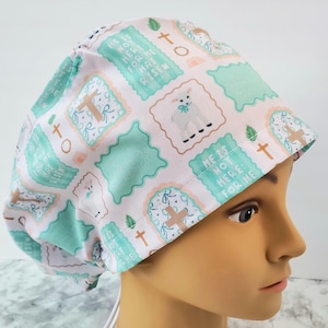 Op de afbeelding: Een witte en groene scrubcap met een herhalend patroon van kruisen, lammeren en de tekst "He is not here: for he has risen." De cap heeft een lichtroze rand en wordt door een persoon gedragen.