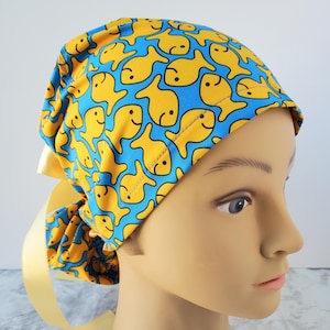 May include: A scrub cap featuring a repeating pattern of yellow goldfish on a blue background. The cap has a yellow ribbon tie and is designed to be worn on the head. The fabric is a bright, cheerful design.