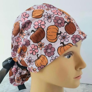 May include: A pink and brown patterned surgical scrub hat with a black tie. The pattern features pumpkins and flowers.