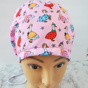 May include: A pink scrub cap featuring a whimsical pattern of cartoon geese dressed in various outfits, including dresses and hats. The geese are interspersed with small white flowers. The cap is designed to be worn in a medical or professional setting.