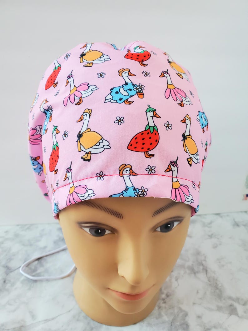 May include: A pink scrub cap featuring a whimsical pattern of cartoon geese dressed in various outfits, including dresses and hats. The geese are interspersed with small white flowers. The cap is designed to be worn in a medical or professional setting.