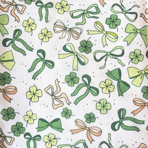May include: A white fabric with a repeating pattern of green, light green, and peach bows and four-leaf clovers. The bows vary in style and size, creating a whimsical and festive design. Small gray dots are scattered throughout the background.