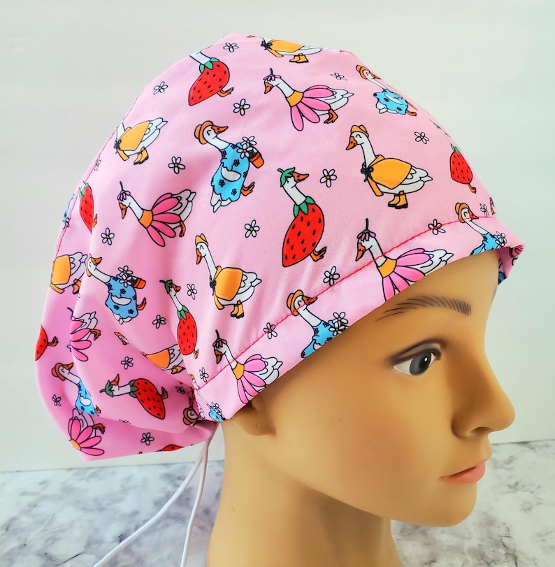 May include: A pink scrub cap featuring a whimsical pattern of cartoon geese in various outfits, strawberries, and small flowers. The cap is made of a soft fabric and has a drawstring for an adjustable fit. The cap is designed for medical professionals.