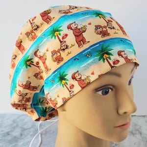 May include: A beige surgical scrub cap featuring a repeating pattern of cartoon monkeys, palm trees, and a beach scene with blue water. The cap has a white drawstring and is designed to cover the head.