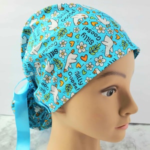 May include: A light blue scrub cap featuring a pattern of white geese, flowers, and orange hearts. The words "Silly Goose!" are printed throughout the design. The cap has a matching blue ribbon tie. very cute