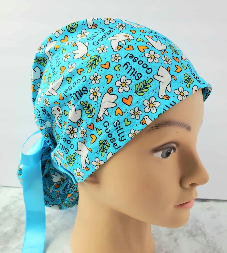 May include: A light blue scrub cap featuring a pattern of white geese, flowers, and orange hearts. The words "Silly Goose!" are printed throughout the design. The cap has a matching blue ribbon tie. very cute