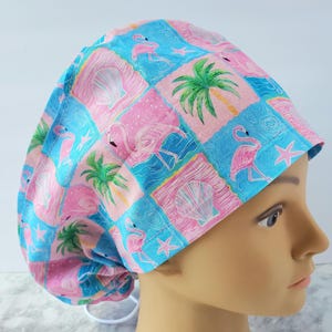 May include: A colorful surgical scrub cap featuring a tropical design. The cap is patterned with pink flamingos, seashells, palm trees, and starfish on a blue and pink checkered background. The cap has a drawstring for a secure fit.