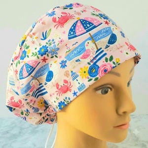 May include: A scrub cap with a light pink background featuring a repeating pattern of beach-themed illustrations including seagulls, blue and pink striped umbrellas, red crabs, lighthouses, seashells, and various colorful flowers.