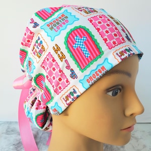 May include: A white scrub cap featuring a colorful pattern of pink checkered squares, blue crosses, and text that reads "Chosen," "Worthy," "Loved," and "Loved by the King." The cap includes a pink ribbon tie at the back for an adjustable fit.