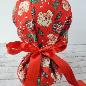 May include: Red fabric scrub hat with a Christmas theme featuring Santa Claus, candy canes, and disco balls. The hat is tied with a red satin ribbon.