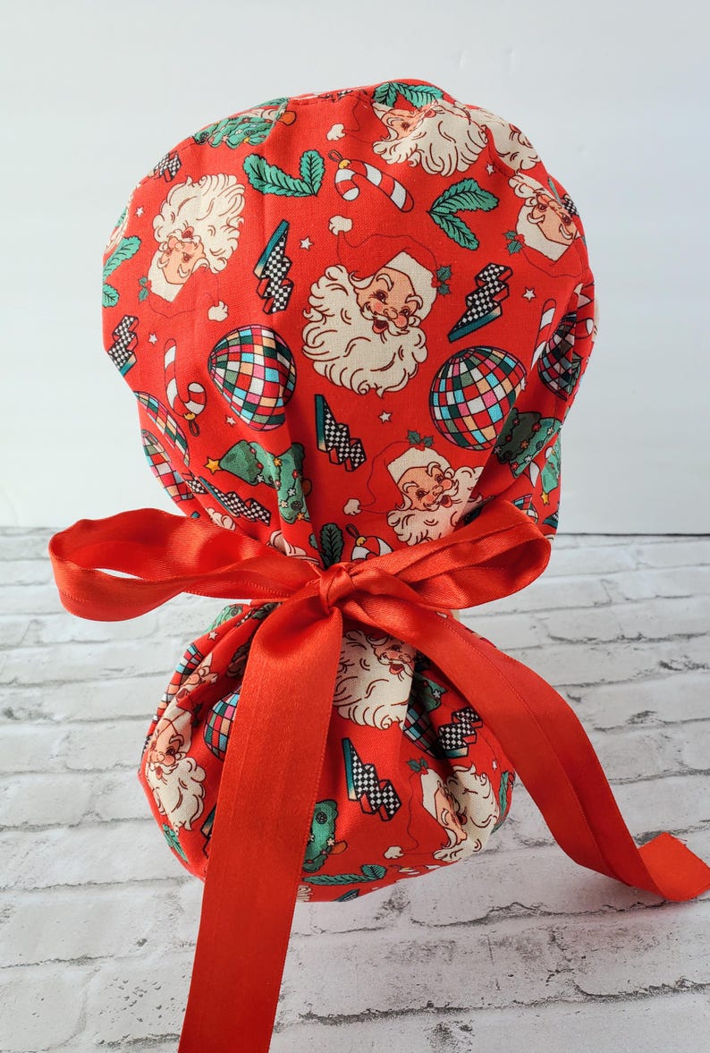 May include: Red fabric scrub hat with a Christmas theme featuring Santa Claus, candy canes, and disco balls. The hat is tied with a red satin ribbon.
