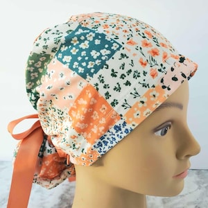 May include: A patchwork scrub cap featuring a floral pattern in shades of orange, white, green, and blue. The cap has a peach-colored ribbon tie. The cap is designed to be worn on the head.