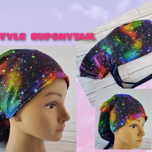 May include: A colorful galaxy print surgical scrub hat with a ponytail opening. The hat is tied with a navy blue ribbon.  The hat is shown on a model with a ponytail.