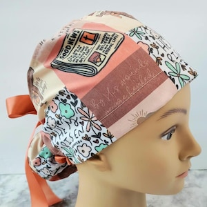 Op de afbeelding: Een roze, beige en witte gepatroneerde chirurgische scrub-muts met een strik. De muts heeft een krantenprint met de woorden "Good News" en een bloemenprint met de woorden "He is risen" en "By His wounds we are healed".