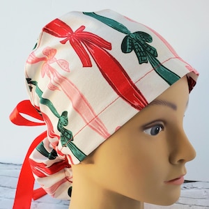 May include: A white scrub hat with a red and green bow pattern. The hat has a red ribbon tie.