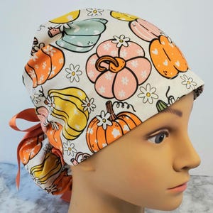 Puede incluir: Un gorro quirúrgico blanco con un estampado de calabazas y flores de colores. Las calabazas son de color naranja, amarillo y rosa, con enredaderas verdes y margaritas blancas. El gorro tiene una cinta color melocotón.