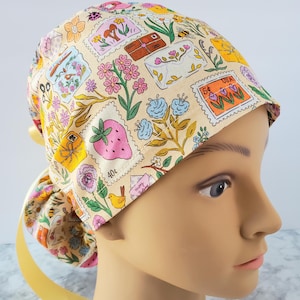 May include: A beige scrub cap with a colorful floral and stamp design. The cap features various illustrations, including a strawberry, flowers, and a bird. A yellow ribbon is tied at the back. This cap is designed for medical professionals.