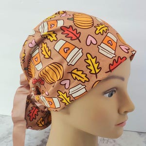 Puede incluir: Un gorro de quirófano marrón con un estampado de temática otoñal. El diseño incluye tazas de café, calabazas y hojas de otoño en naranja, amarillo y blanco. El gorro tiene un lazo de cinta de satén a juego.