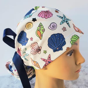 May include: A white scrub hat with a colorful seashell and starfish pattern. The hat has a navy blue tie.