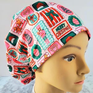 May include: A scrub cap featuring a light pink background with a repeating pattern of Christmas-themed icons in scalloped frames, including nutcrackers, gingerbread men, candy canes, gift boxes, and the text Merry Christmas in red letters.