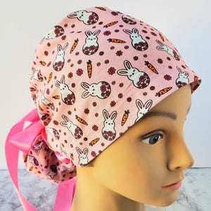 Könnte beinhalten: Eine rosa OP-Haube mit einem sich wiederholenden Muster aus weißen Hasen mit floralen Akzenten, orangefarbenen Karotten und kleinen Blumen. Die Haube hat hinten eine rosa Schleife. Für medizinisches Fachpersonal.
