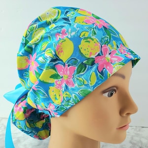 Puede incluir: Un gorro quirúrgico azul con un estampado de limones amarillos y flores rosas. El gorro tiene una cinta azul claro para atar en la parte posterior. La tela tiene un diseño brillante y colorido.