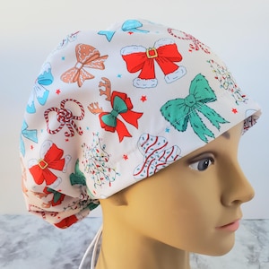May include: White surgical scrub cap with a festive Christmas bow pattern. The cap features various bows in red, green, and blue, along with reindeer and Christmas tree designs. The cap is tied in the back with white ties.