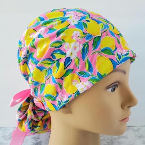 Puede incluir: Un gorro quirúrgico rosa con un estampado vibrante de limones amarillos, flores blancas y hojas azules. El gorro tiene un diseño con lazo en la parte trasera y está hecho de una tela suave estampada. El gorro está diseñado para ser usado en la cabeza.