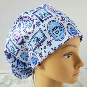 May include: A light blue scrub cap featuring a winter-themed pattern. The design includes framed illustrations of a snowman, ice skates, a mug of hot cocoa, and a cozy sweater. Accents include snowflakes, bows, and flowers.