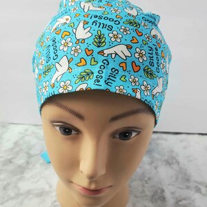 May include: A light blue scrub cap featuring a pattern of white cartoon geese, flowers, hearts, and the words "Silly Goose!" The cap is designed for medical professionals and is made of a soft, breathable fabric.