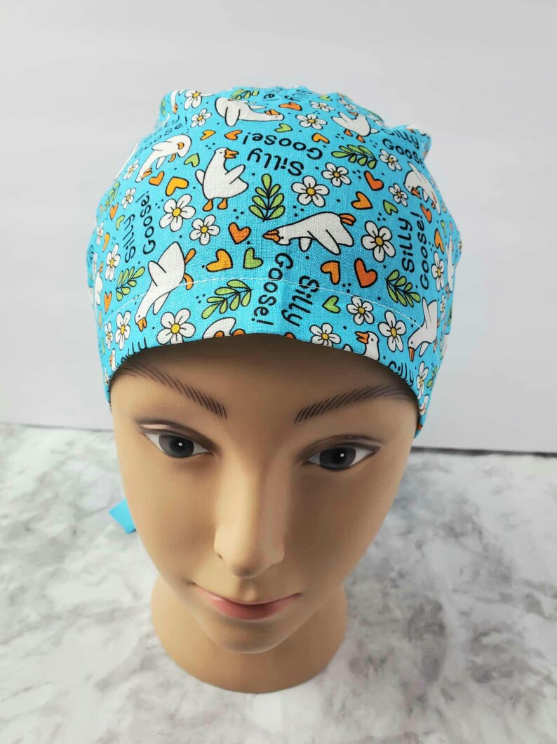 May include: A light blue scrub cap featuring a pattern of white cartoon geese, flowers, hearts, and the words "Silly Goose!" The cap is designed for medical professionals and is made of a soft, breathable fabric.