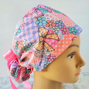Puede incluir: Un gorro quirúrgico colorido con un diseño de patchwork con motivos florales, lazos y estrellas en tonos rosa, naranja, azul y morado. El gorro tiene una cinta rosa en la parte trasera.