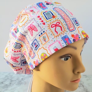 May include: A white scrub cap with a colourful pattern of medical illustrations, including X-ray images, bows, and flowers. The cap features the word "RADIOLOGY" in pink lettering and has a tie-back closure.