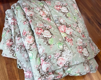 May include: A quilted comforter featuring a sage green background with a repeating floral pattern of pink, white, and cream roses, along with green leaves and small red berries. The fabric has a soft, padded texture with visible stitched lines.