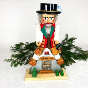 May include: A wooden Swiss man nutcracker figurine. The figurine wears a black top hat, white shirt, red vest, and brown pants. The base features the text "HOLADI...HO" and "THE SWISS MAN". The figurine is 12 inches tall.