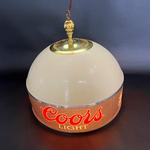 May include: A dome-shaped hanging pendant light fixture featuring a cream-colored top and a textured brown band around the base. The band displays the red and gold Coors Light logo. A polished brass-colored metal cap sits at the very top of the dome.