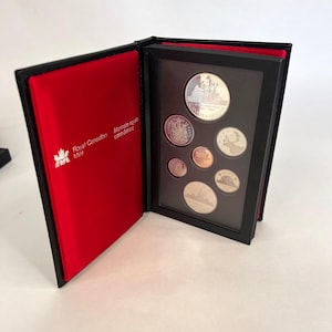 May include: A Royal Canadian Mint coin set displayed in a black book-style case with a red interior lining. The set includes six silver-toned coins of varying sizes, featuring Canadian symbols and text, secured within a black frame inside the case.