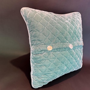 May include: A teal quilted pillow with a soft, velvety texture. The pillow features a diamond pattern and is adorned with two round, off-white buttons. The pillow is a square shape.