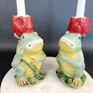 May include: A pair of ceramic candle holders shaped like frogs, each featuring a light green and yellow body with a red lotus flower on its head. The frogs are depicted in a seated position, with white taper candles inserted into the flower tops.