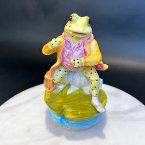 May include: A whimsical ceramic frog figurine, dressed in a yellow coat and pink shirt, holding a basket. The frog is seated on a lily pad base with blue accents. The figurine is a decorative item.