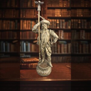 May include: A silver-toned metal figurine of a musketeer standing on a circular base. The figure wears a wide-brimmed hat and period clothing, holding a tall, thin pole with a small decorative top piece that extends above the head.