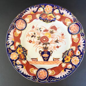 May include: A decorative ceramic plate with a floral design. The plate features a white center with a bouquet of flowers in a blue vase. The rim is decorated with a blue, red, and gold pattern. The plate measures approximately 10 inches in diameter.
