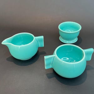 May include: Three teal ceramic bowls, two with handles, one with a rounded base. The bowls are all the same color and have a smooth, glossy finish.