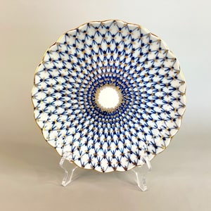 May include: A white porcelain plate with a blue and gold net pattern. The plate has a scalloped edge with gold trim. The intricate design radiates from the center, creating a decorative and elegant aesthetic. The plate is displayed on a clear stand.