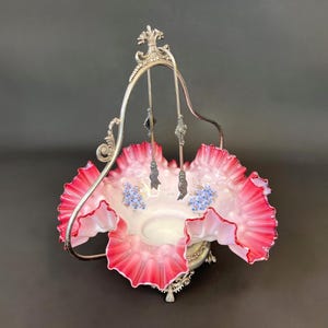 May include: An antique silver-plated serving basket with a ruffled, pink-edged glass bowl. The bowl has a white interior and is decorated with blue floral accents. The basket has a decorative handle and hanging serving utensils.