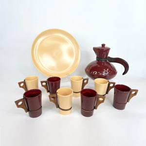May include: A vintage coffee set featuring a large, round, cream-colored plate, a maroon coffee pot with a wooden handle, and eight small coffee cups. The cups alternate between cream and maroon, each with a wooden handle.