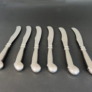 May include: Set of seven silver butter knives. Each knife has a curved blade and a decorative handle with a swirl design at the base. The knives are arranged on a dark surface.